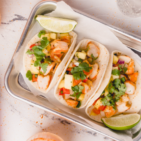 Three shrimp tacos with pineapple and cilantro on a metal tray.