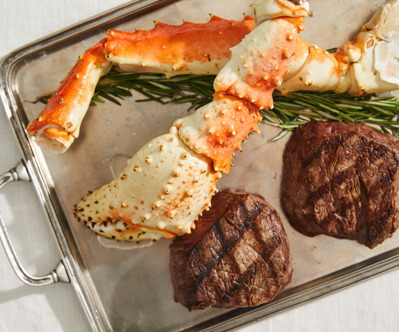 Crab legs and grilled steaks on a metal tray with rosemary.