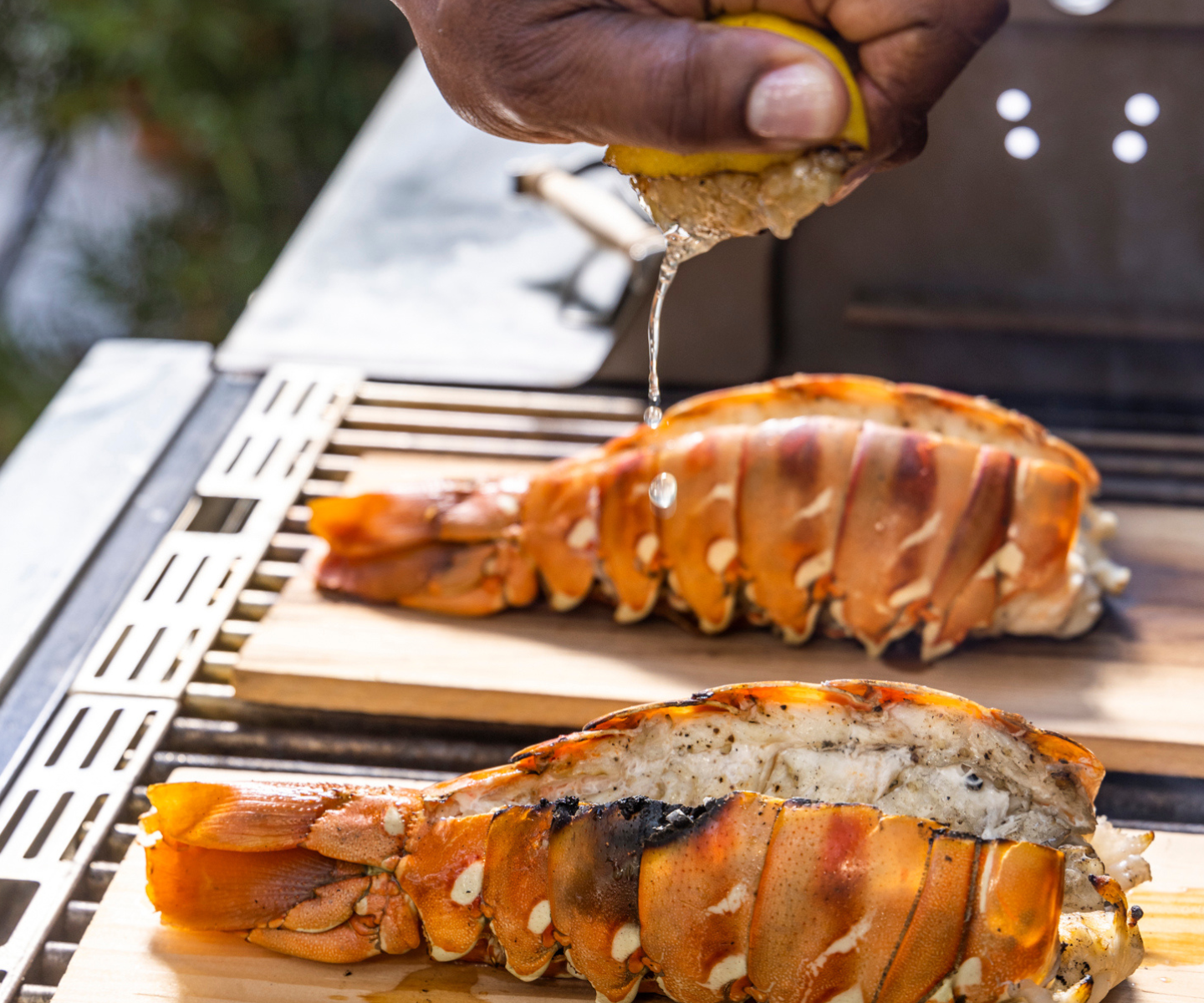 Grilled lobster tails on a cutting board with a hand squeezing lemon over them.