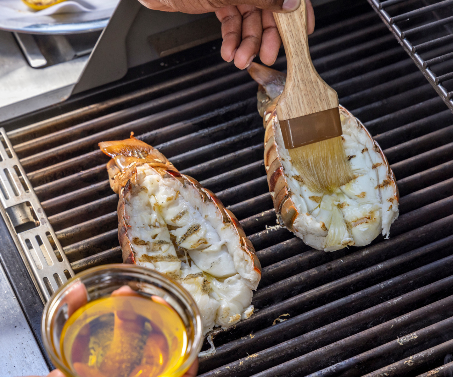 Lobster tails being grilled with a brush and a small bowl of butter on a grill.