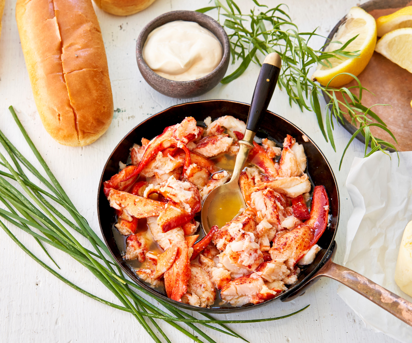Lobster dish with a spoon in a skillet on a white surface with bread and lemon slices.