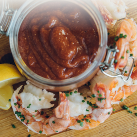 Jar of cocktail sauce on a wooden board with shrimp and lemon