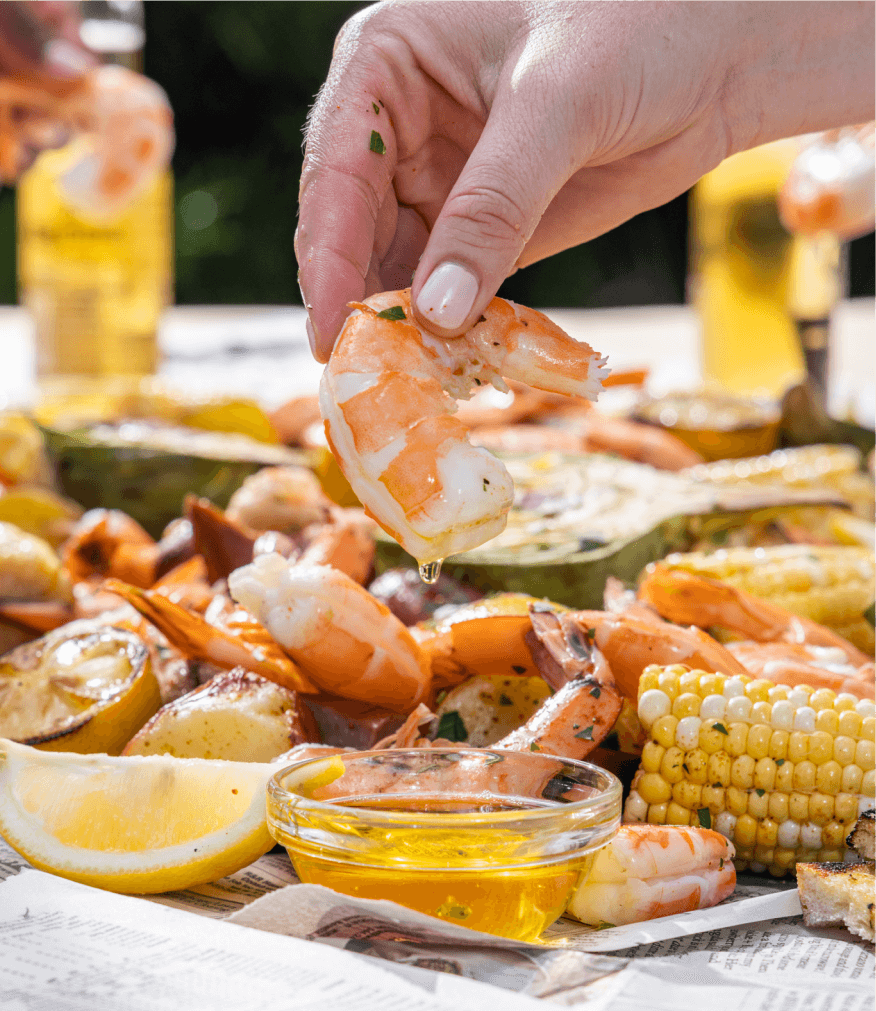hand dipping shrimp into butter on table with corn, potatoes, lemons and other crab boil fixings.