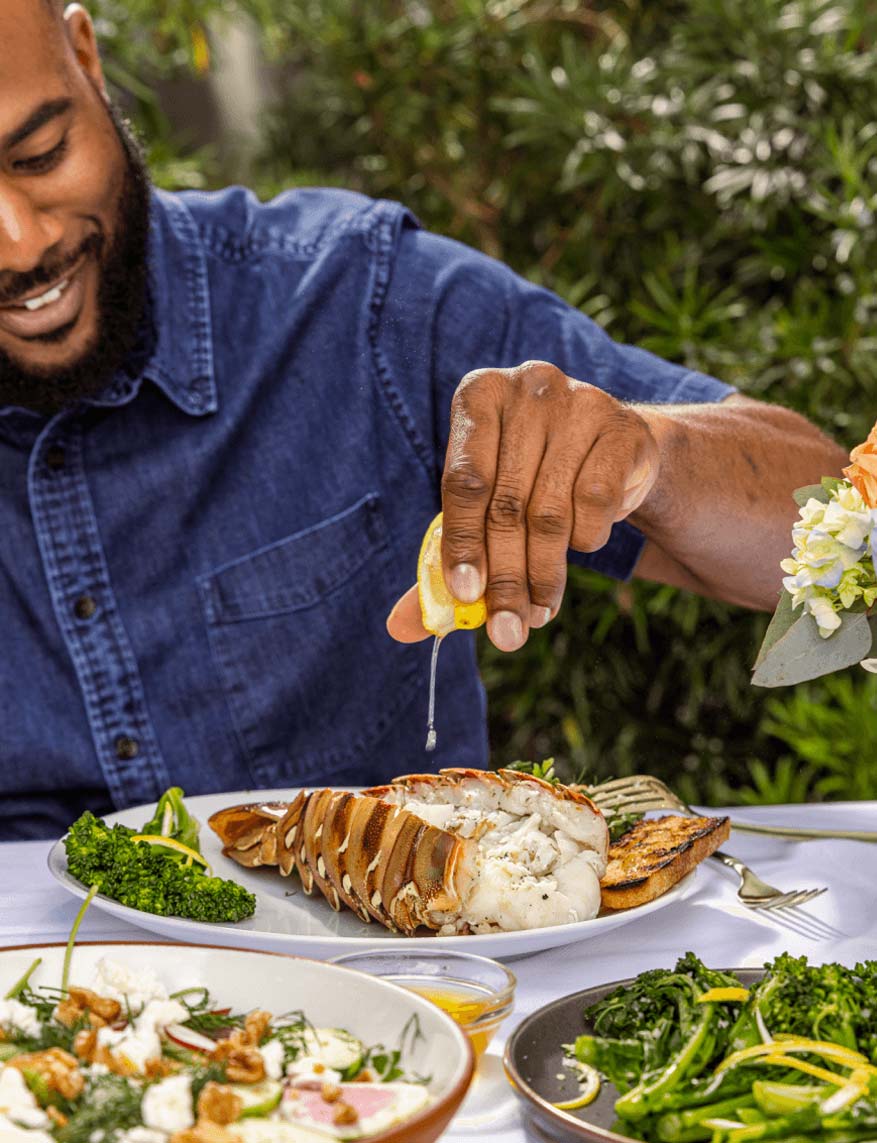 Man squeezing lemon juice over lobster tail at an outdoor table.