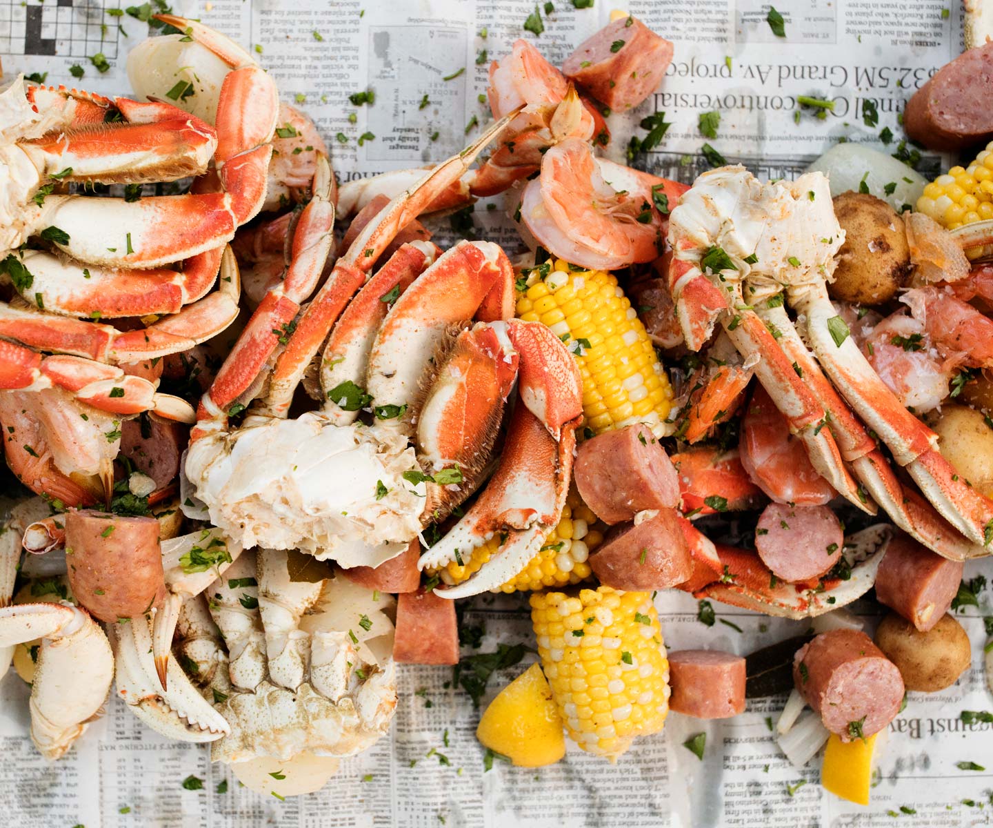 Plated seafood dish with crab legs, corn on the cob, and hot dogs on a newspaper background