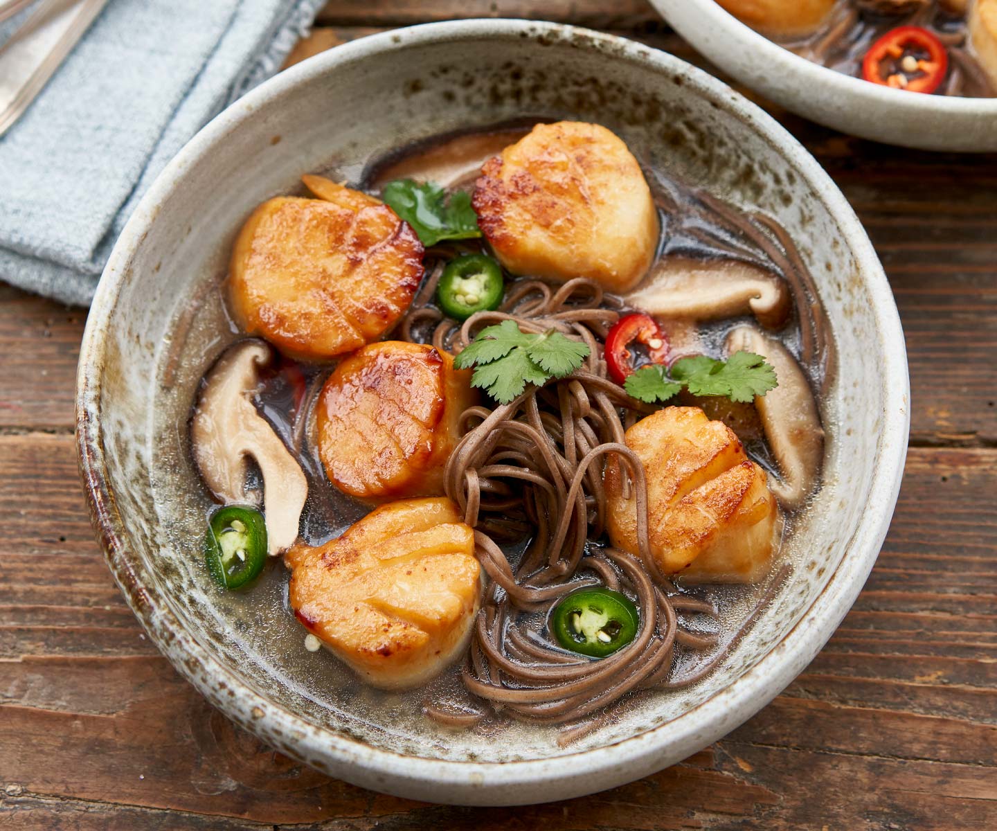 Bowl of soup with scallops, noodles, and vegetables on a wooden table
