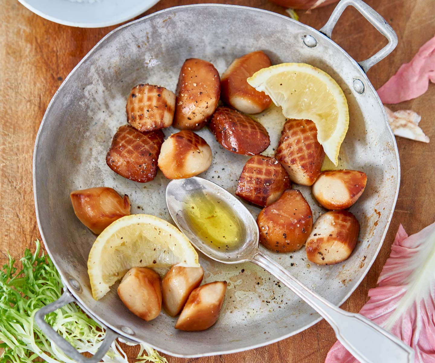 Scallops in a pan with lemon wedges and a spoon on a wooden surface