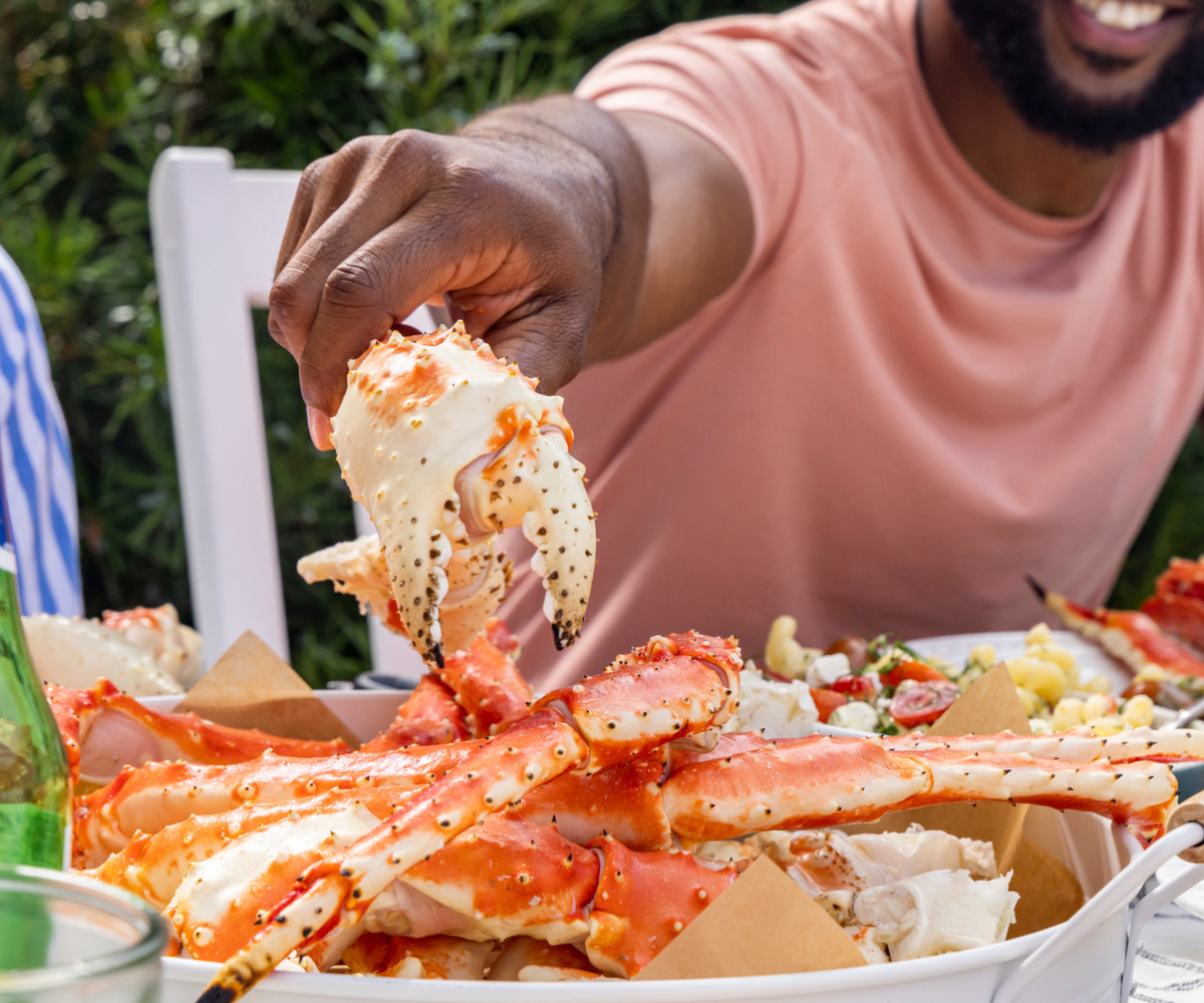 Person holding a crab leg over a plate of crab and vegetables outdoors.