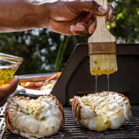 Person brushing garlic butter on grilled lobster tails with a wooden brush.