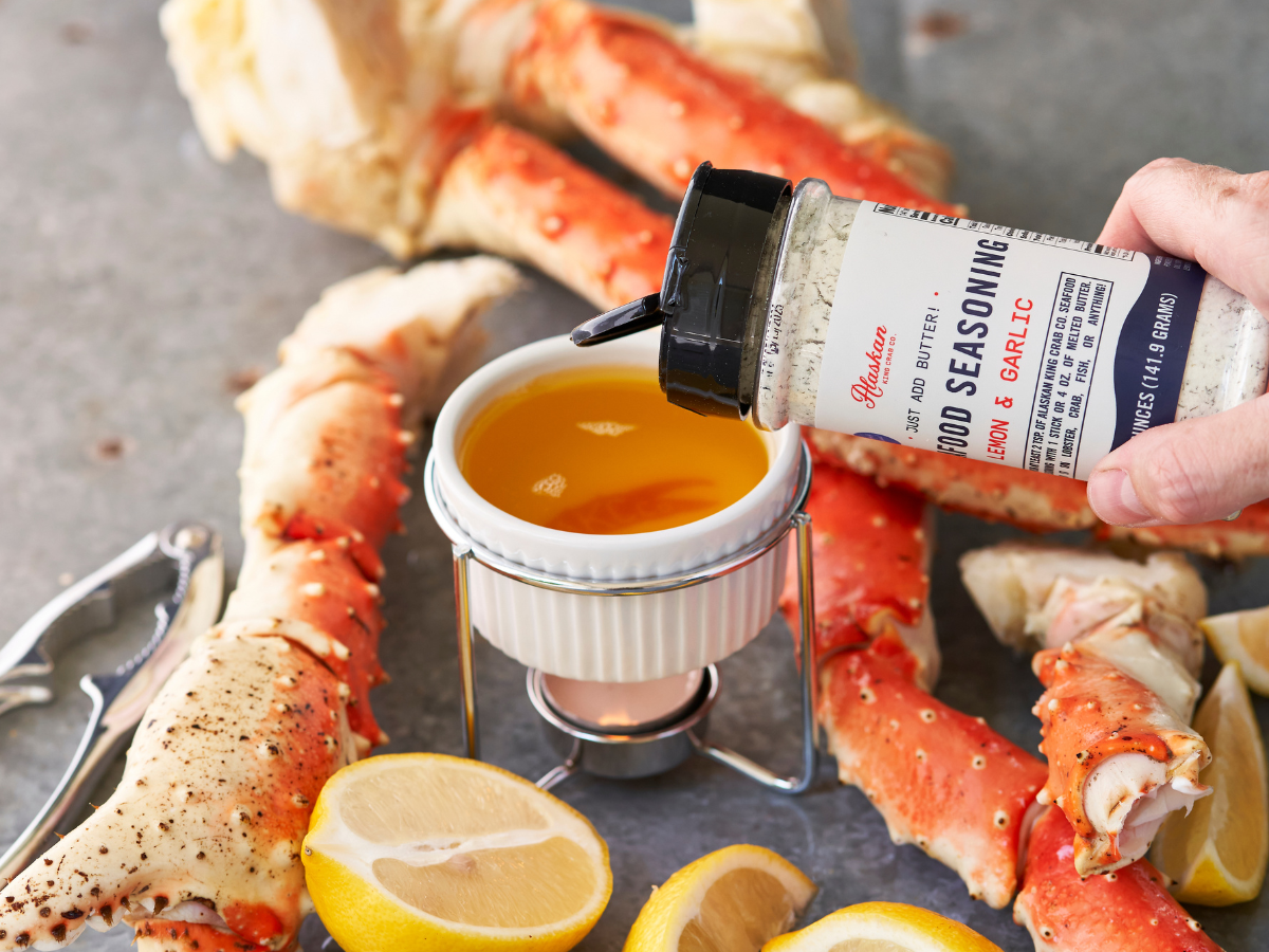 Person seasoning crab legs with a container of Gander seasoning on a metal surface.