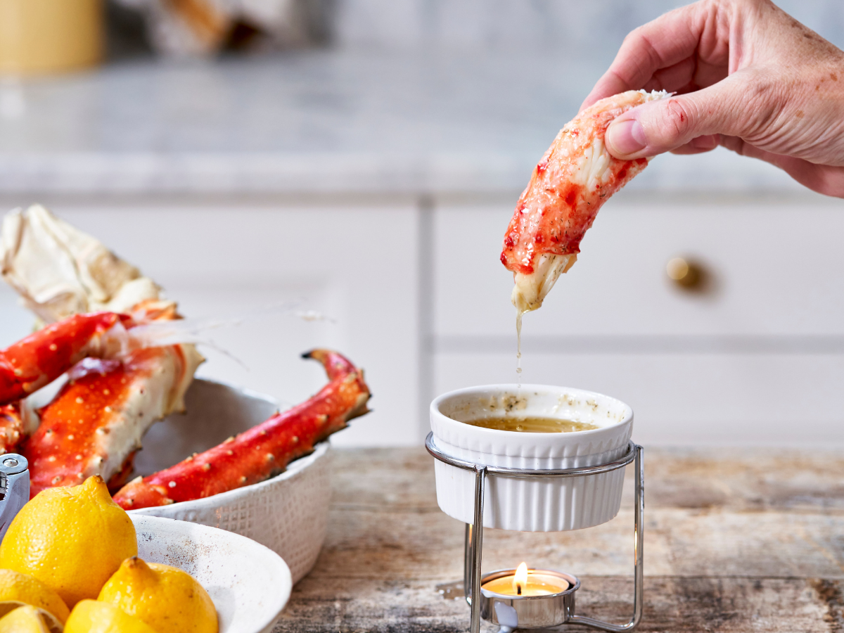 Person dipping a crab leg into a small bowl of sauce with lemons and more crab legs in the background.