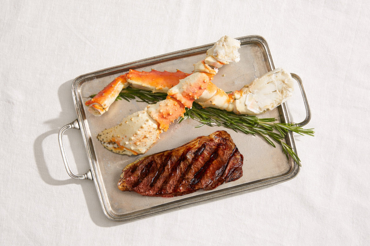 Steak and crab legs on a metal tray with rosemary on a white background