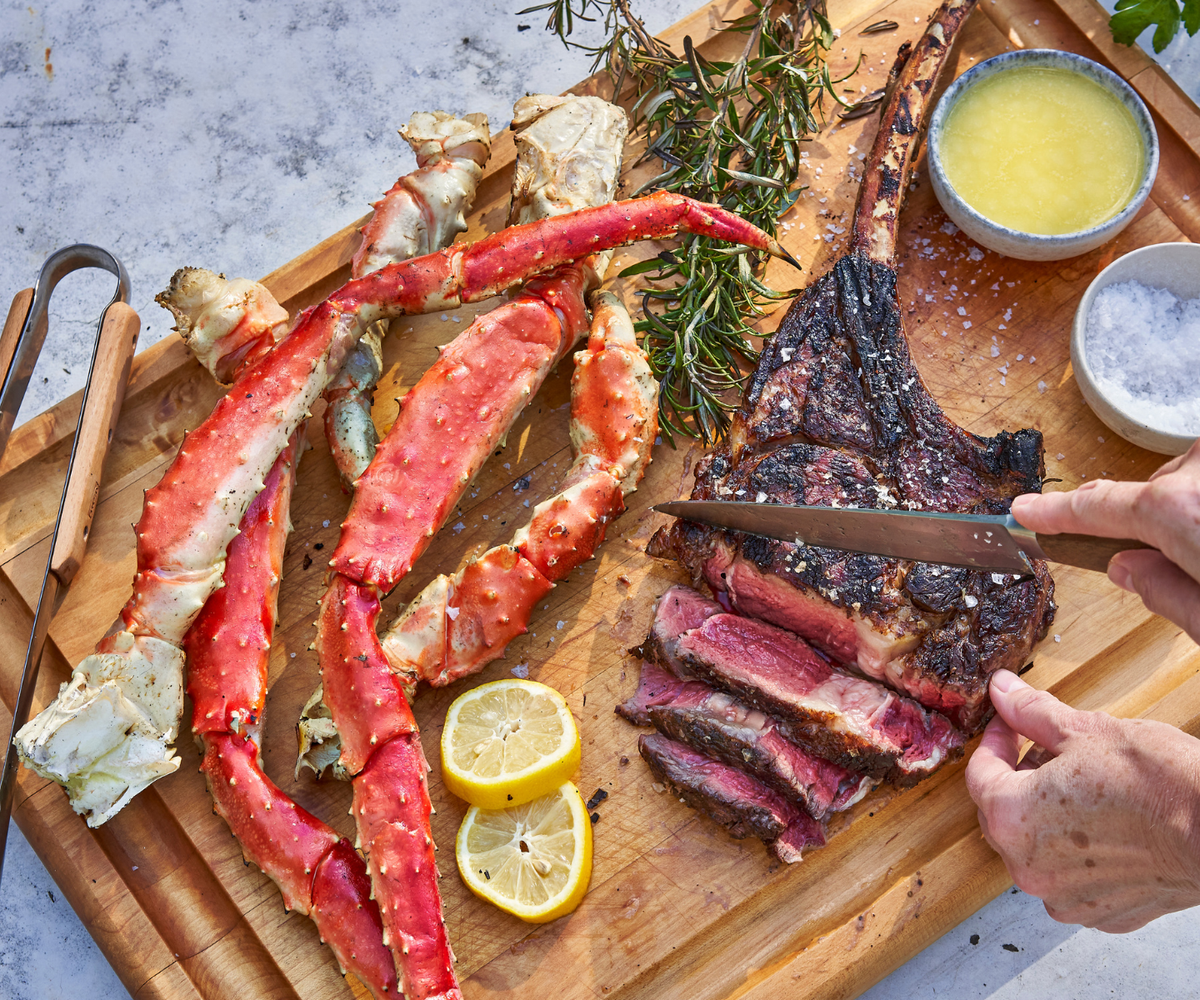 Wooden cutting board with cooked steak, crab legs, and lemon wedges on a light background