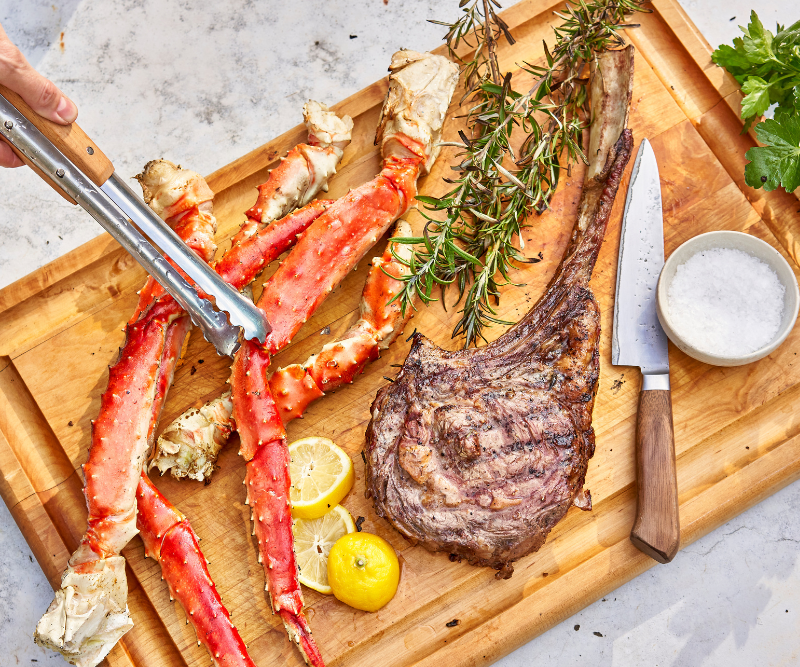 Wooden cutting board with cooked crab legs, steak, lemon wedges, and herbs on a light background.