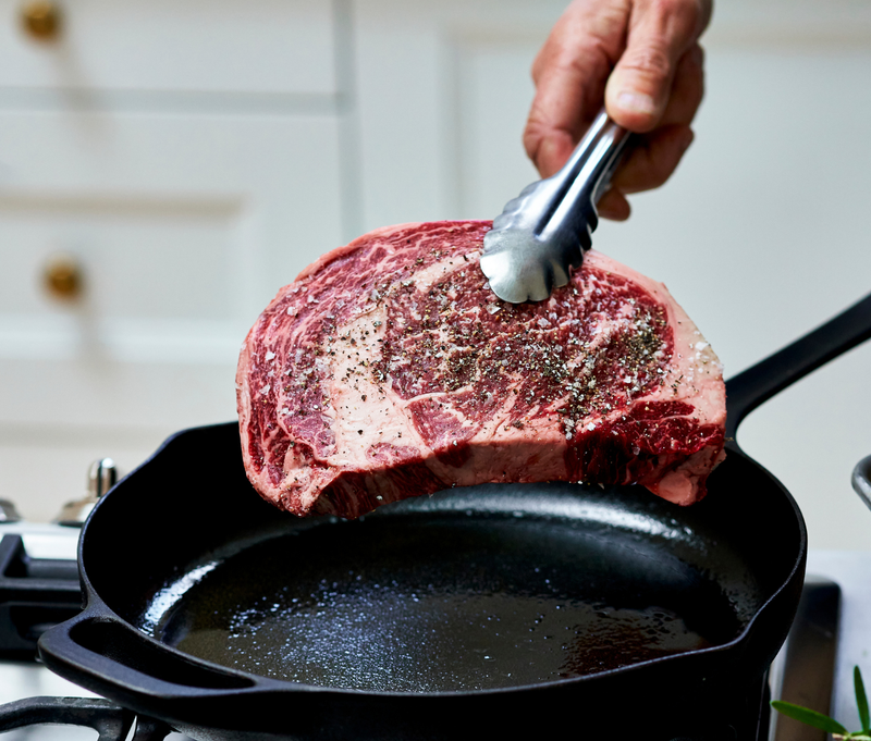 Raw steak being placed into a hot cast iron skillet with a blurred kitchen background