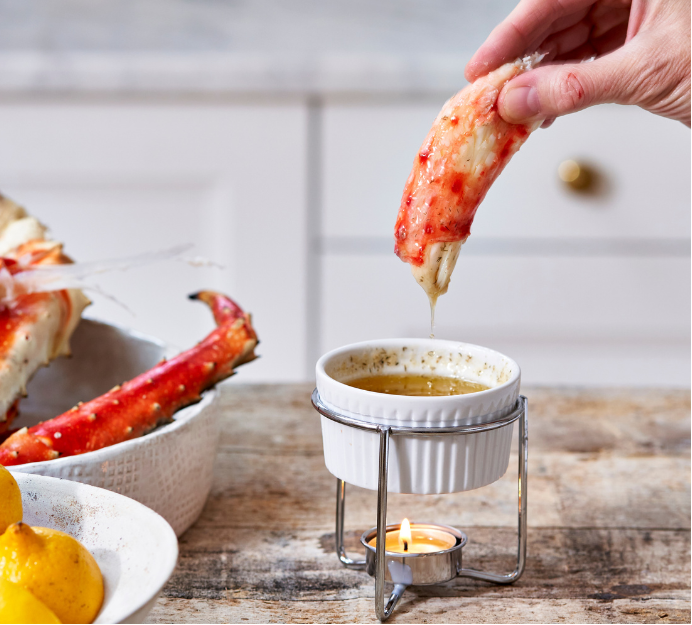 Person dipping a crab leg into a small bowl of butter on a wooden table with lemons and more crab legs in the background.