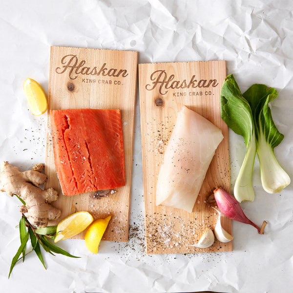 Two wooden cutting boards with Alaskan King Crab Co branding, featuring raw fish and vegetables on a white background.