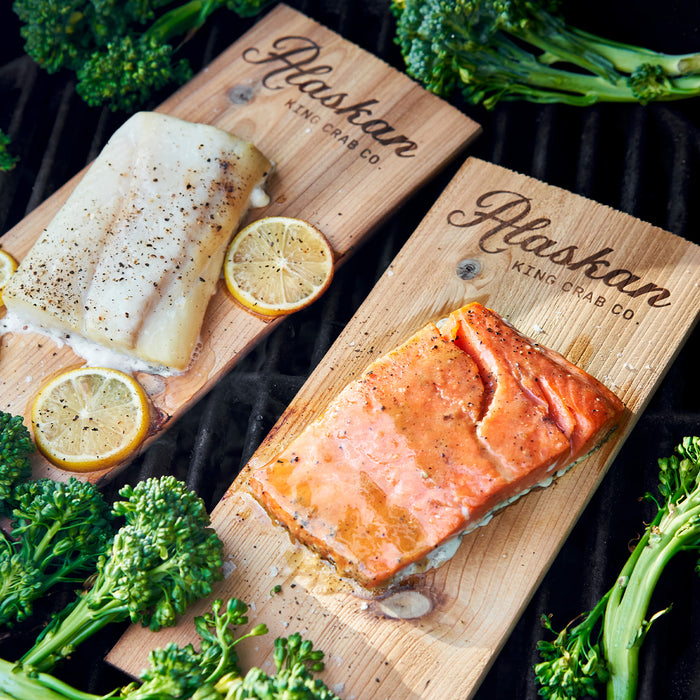 Two wooden cutting boards with salmon and cod fillets, garnished with lemon slices and surrounded by broccoli, on a dark background.