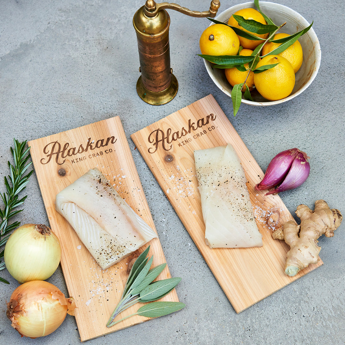 Two pieces of fish on wooden boards with 'Alaskan King Crab Co.' branding, surrounded by lemons, onions, and a pepper grinder.