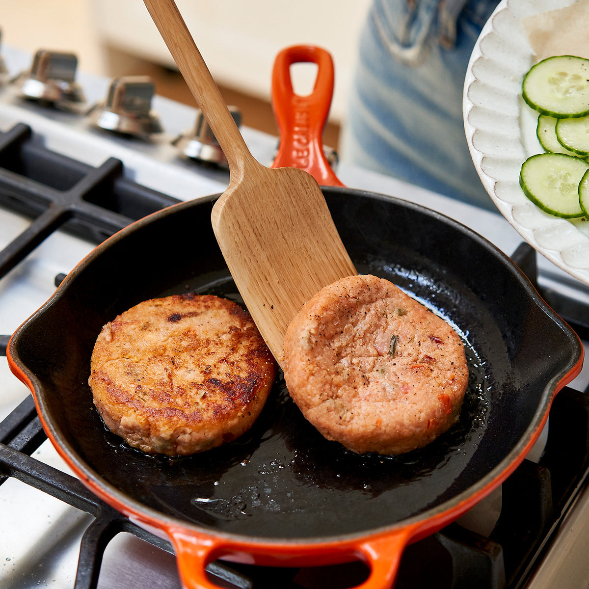 Two salmon patties cooking in a black skillet with a wooden spatula on a stove.