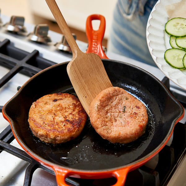 Two salmon patties cooking in a black skillet with a wooden spatula on a stove.