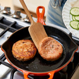Two salmon patties cooking in a black skillet with a wooden spatula on a stove.