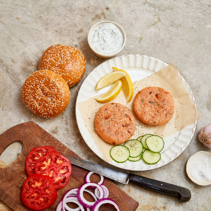 Salmon burgers on a plate with buns, vegetables, and a knife on a stone surface.