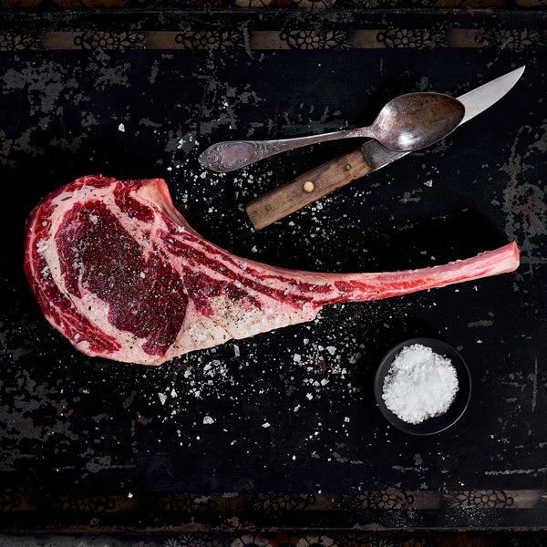Uncooked tomahawk ribeye seasoned everywhere with salt and is lying by a spoon and knife.