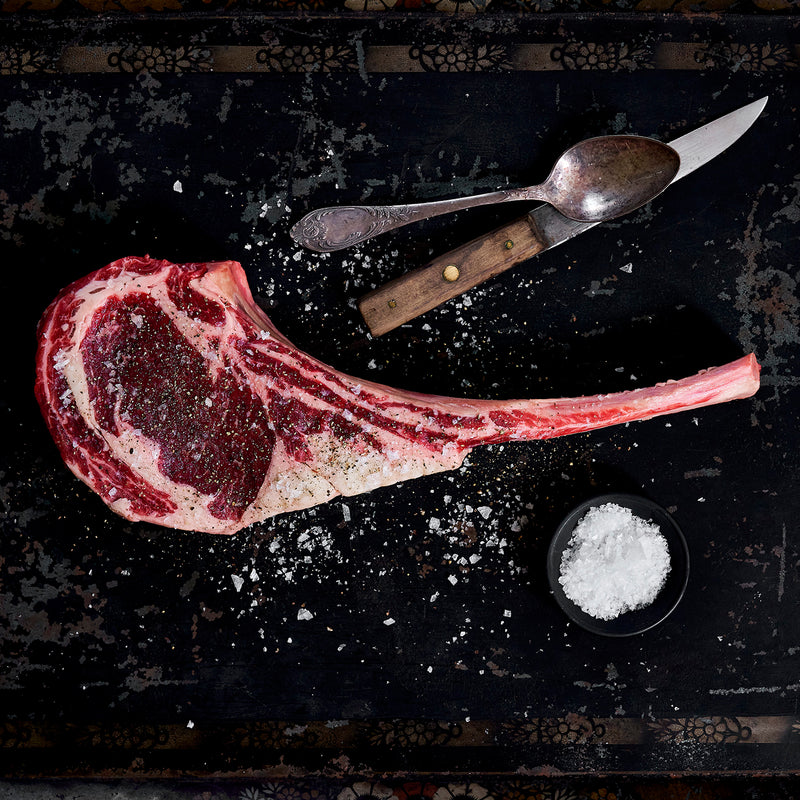 Uncooked tomahawk ribeye seasoned everywhere with salt and is lying by a spoon and knife.