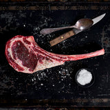 Uncooked tomahawk ribeye seasoned everywhere with salt and is lying by a spoon and knife.