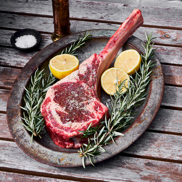 Uncooked tomahawk ribeye on a metal plate with lemons cut in halves and rosemary. 