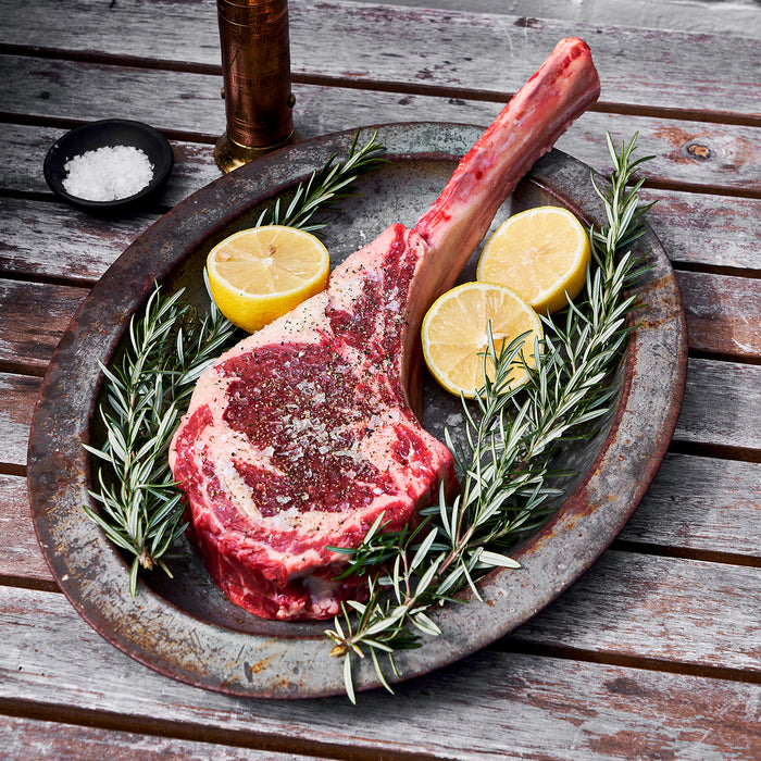 Uncooked tomahawk ribeye on a metal plate with lemons cut in halves and rosemary. 