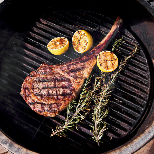 Tomahawk steak on the grill with rosemary and lemons cut in half.