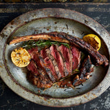 Cooked tomahawk ribeye with lemon and rosemary on metal plate. The ribeye is being served off the bone. 