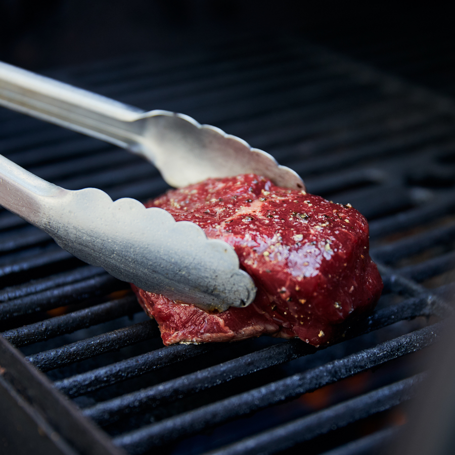 Raw steak being held by tongs over a grill