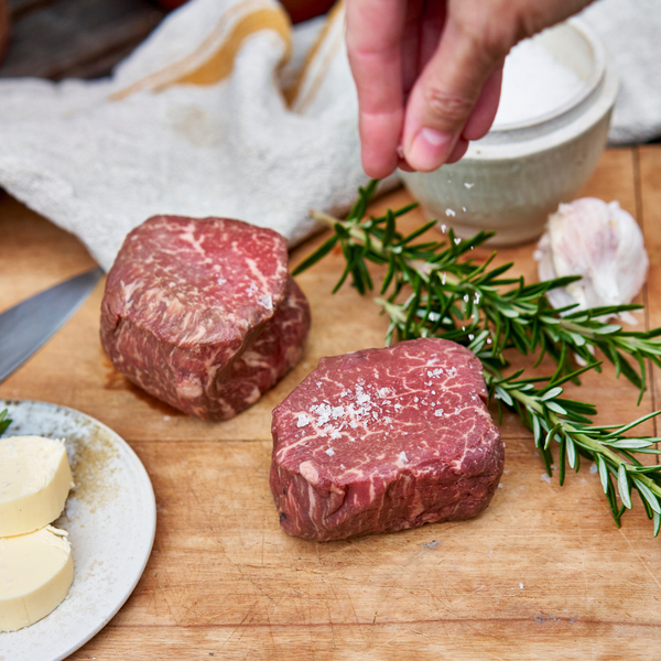 Raw beef steaks on a wooden cutting board with rosemary and a hand sprinkling salt.