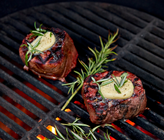Two steaks on a grill with butter and rosemary, surrounded by flames.
