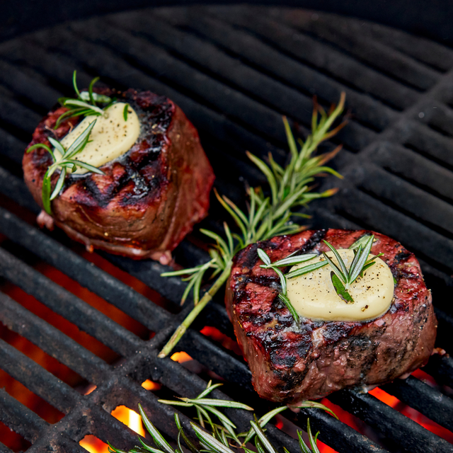Two steaks on a grill with butter and rosemary, surrounded by flames.