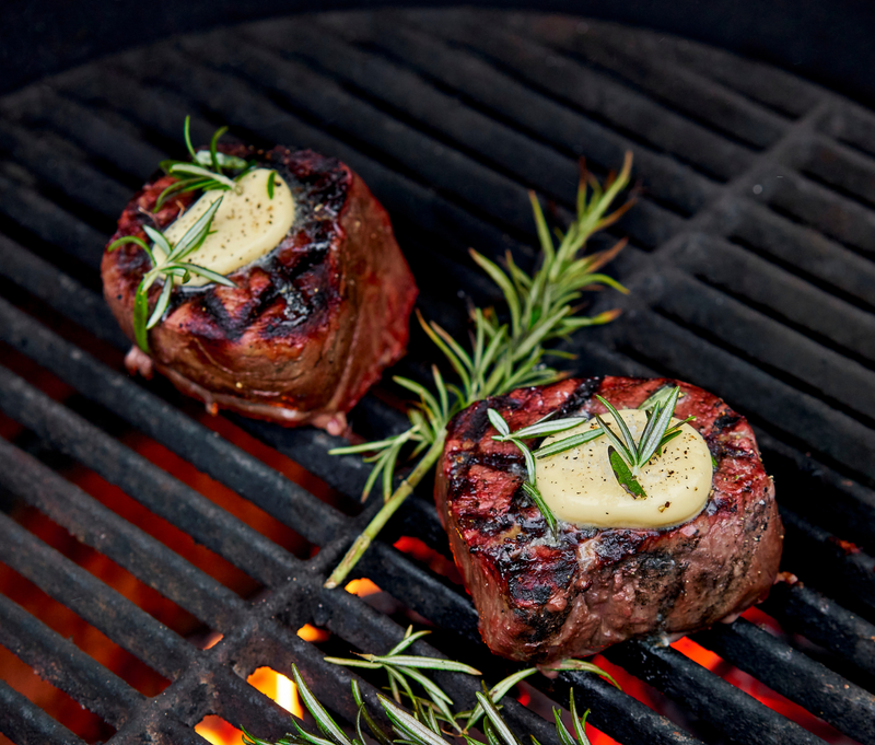 Two steaks on a grill with butter and rosemary, surrounded by flames.