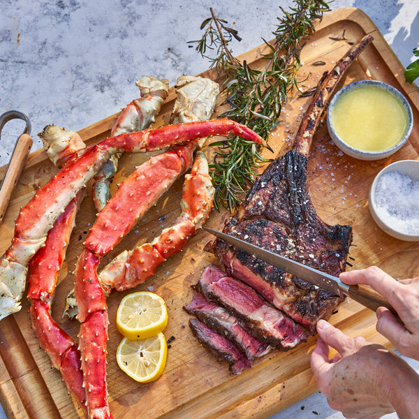 Wooden cutting board with grilled steak, crab legs, and lemon slices on a marble surface.