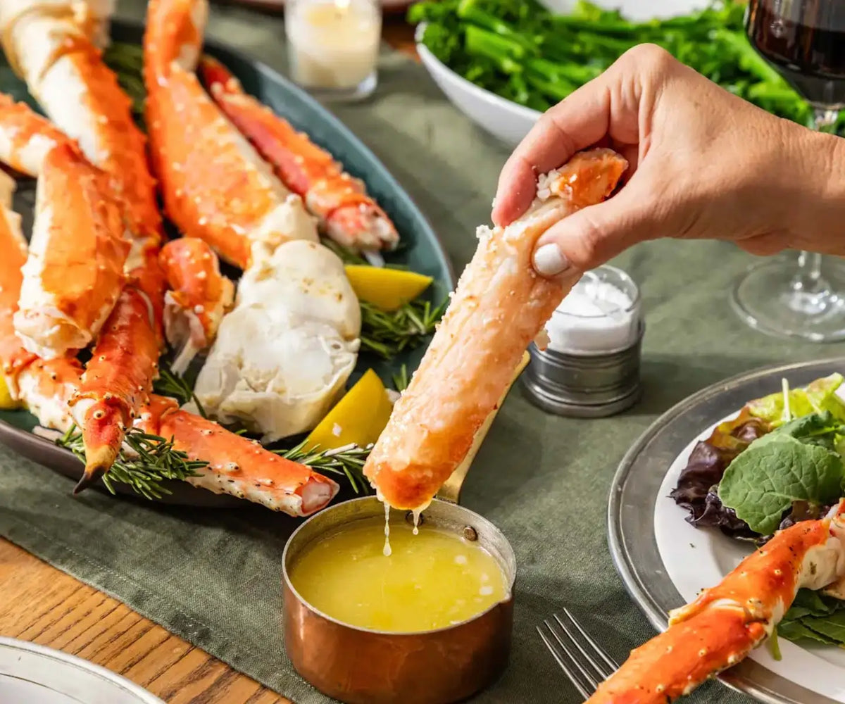 Person dipping a crab leg into a bowl of butter on a dining table with seafood and salad.