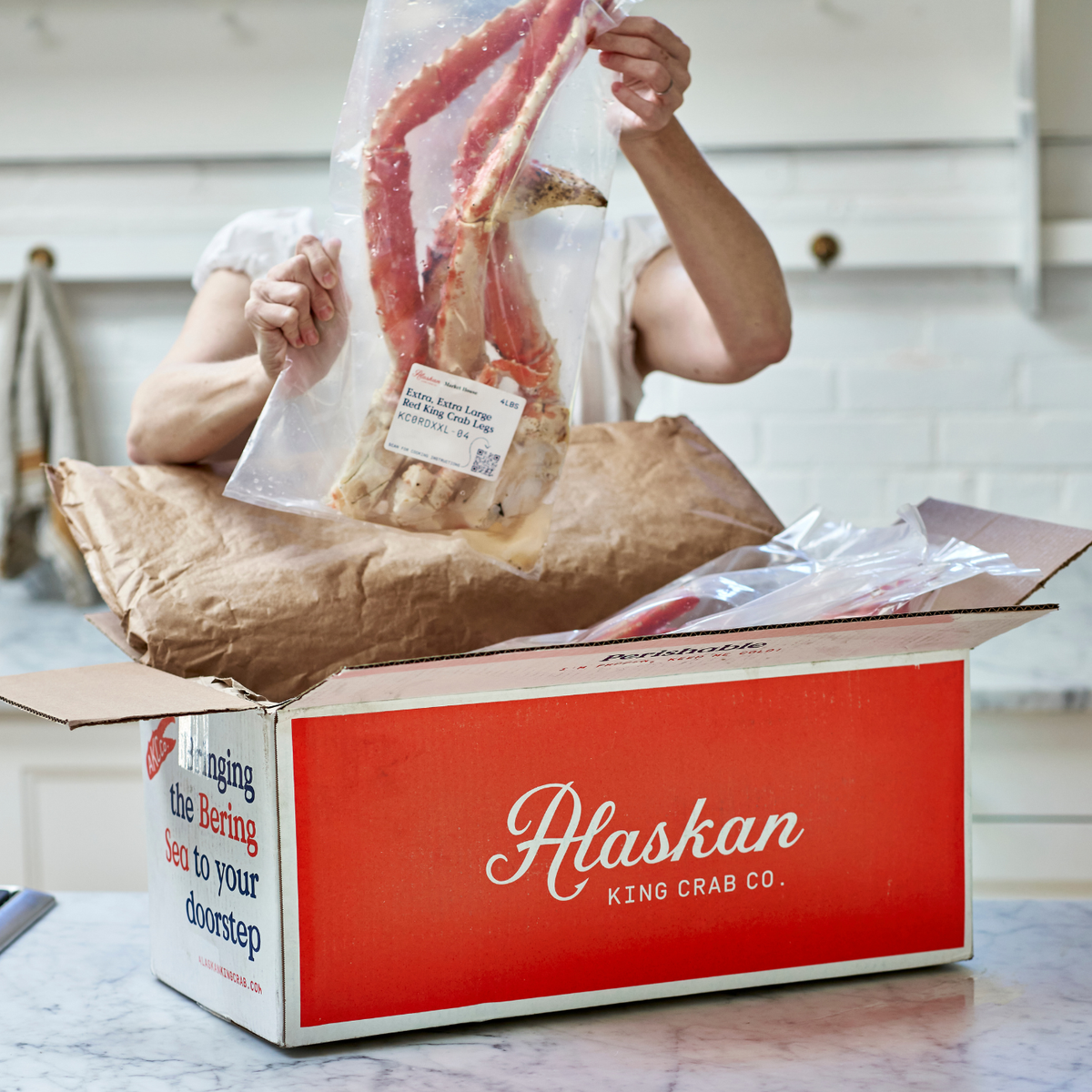 Person unpacking Alaskan King Crab Co. box with king crab legs and other seafood in a kitchen.